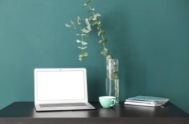 Laptoplu iş yeri, vazoda okaliptüs, kupa ve yeşil duvarın yanındaki defterler.