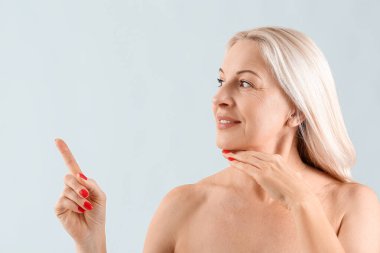 Sağlıklı ciltli olgun sarışın kadın açık arkaplanda bir şeyi işaret ediyor.