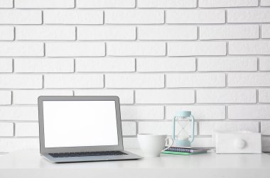 Beyaz tuğla duvarın yanında dizüstü bilgisayarı, fincanı ve kum saati olan işyeri