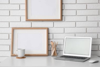 Diz üstü bilgisayarı, çerçeveleri ve beyaz tuğla duvarın yanındaki bardağı olan işyeri