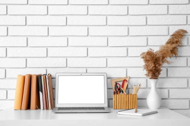 Beyaz tuğla duvarın yanındaki vazoda dizüstü bilgisayarı, kırtasiyesi ve pampası olan işyeri.