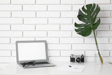 Beyaz tuğla duvarın yanındaki vazoda dizüstü bilgisayarı, fotoğraf makinesi ve palmiye yaprağı olan işyeri.