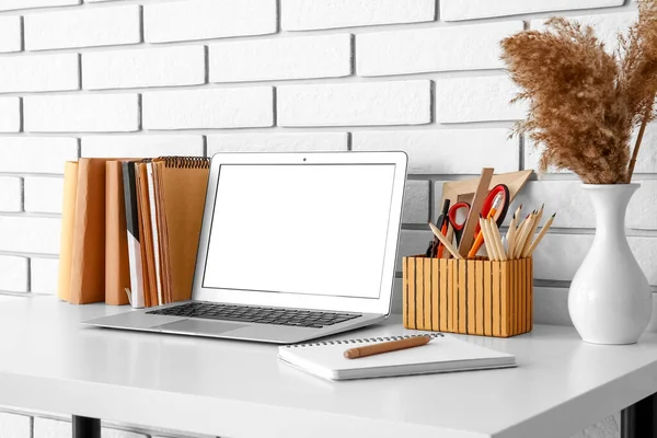 Beyaz tuğla duvarın yanındaki vazoda dizüstü bilgisayarı, kırtasiyesi ve pampası olan işyeri.