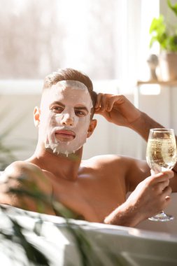 Yüzünde maske ve bir bardak şarapla evde banyo yapan yakışıklı bir adam.