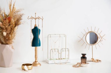 Stands with stylish jewelry, vase with flowers, mirror and perfume bottle on chest of drawers near light wall