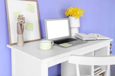 Laptop, bardak, defter ve vazo renkli duvarın yanında çiçekler.