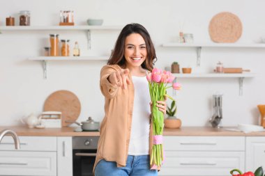 Çiçek buketi olan genç bir kadın, izleyiciyi evine davet ediyor.