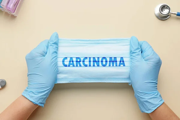 Hands of doctor holding medical mask with word CARCINOMA on light background, top view