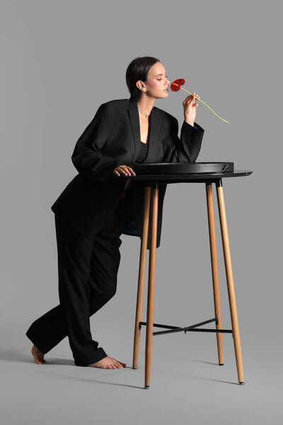 Elegant woman standing near modern table and smelling anthurium flower on grey background