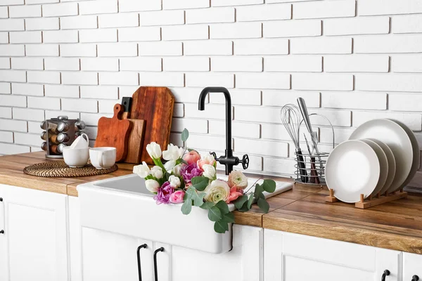 Güzel çiçeklerle lavabo ve mutfak gereçleri beyaz tuğla duvarın yanındaki tezgahlarda.