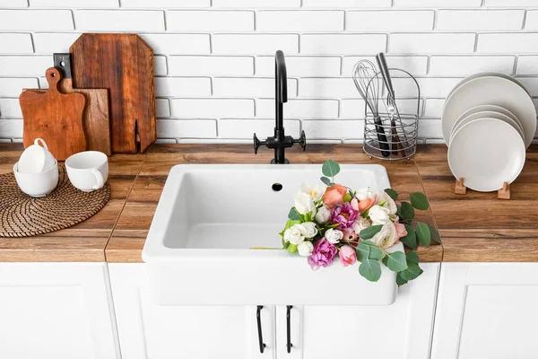 Güzel çiçeklerle lavabo ve mutfak gereçleri beyaz tuğla duvarın yanındaki tezgahlarda.