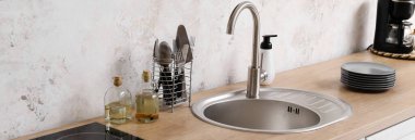 Gümüş lavabolu ahşap tezgah, farklı çatal bıçaklar ve mutfak duvarının yanındaki yağ şişeleri.