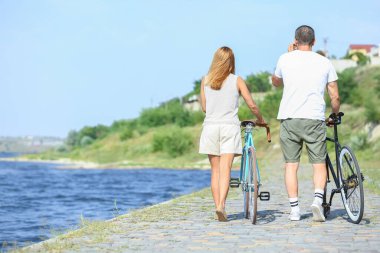 Yaz günü nehir kıyısında bisiklet süren olgun bir çift.