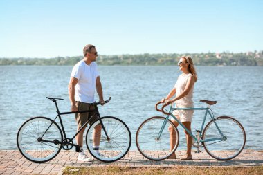 Yaz günü nehir kıyısında bisiklet süren olgun bir çift.