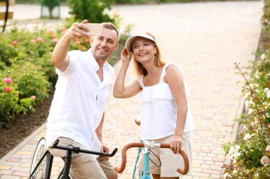 Bisikletli olgun bir çift yaz günü şehir parkında selfie çekiyor.