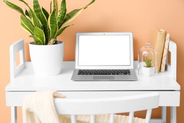 Boş ekranlı dizüstü bilgisayar, kitaplar ve bej duvarın yanındaki masada bitkiler.