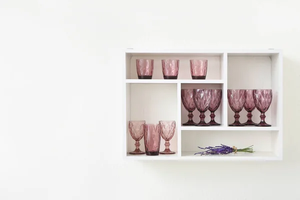 Shelf unit with empty stylish glasses and bouquet of lavender flowers on white wall