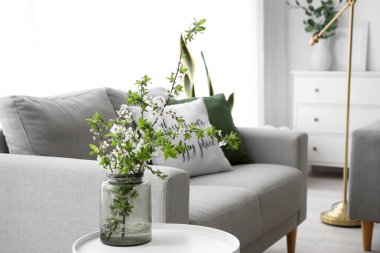 Vase with blooming tree branches on table near sofa in living room