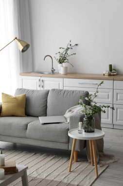 Vase with blooming tree branches and sofa with modern laptop in light room interior