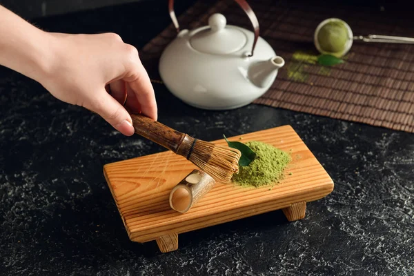 Koyu arka planda Chasen ve pudralanmış kibrit çayla kadın eli, yakın plan.