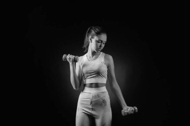 Black and white portrait of beautiful sporty young woman with dumbbells on dark background