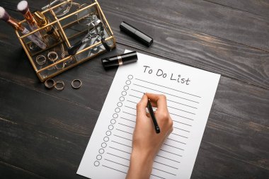 Karanlık ahşap arka planda yapılacaklar listesi, kozmetik ve aksesuarları olan kadın eli, yakın plan