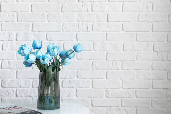 Vase with blue tulips and magazine on table near white brick wall