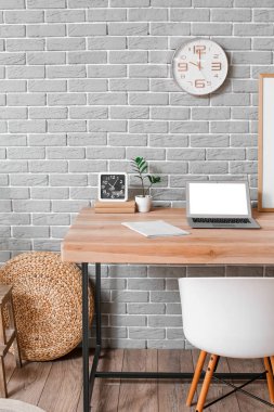Modern dizüstü bilgisayar, defter ve alarm saati gri tuğla duvarın yanında duran ahşap masa üzerinde.
