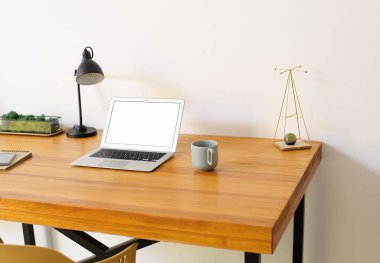 Modern laptop, mobile phone and cup on wooden standing desk near light wall