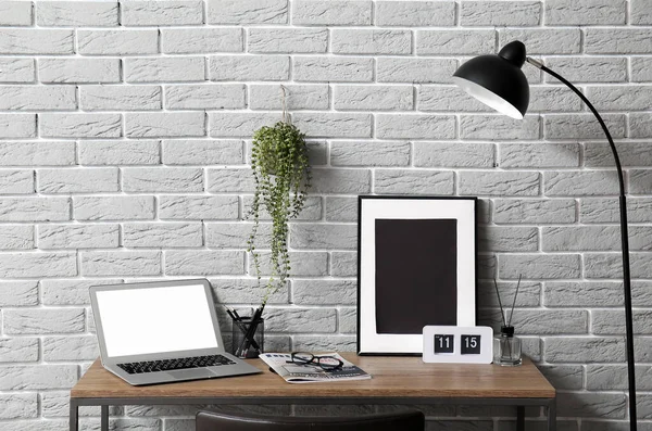 Modern laptop, alarm saati ve resim çerçevesi gri tuğla duvarın yanındaki masada.