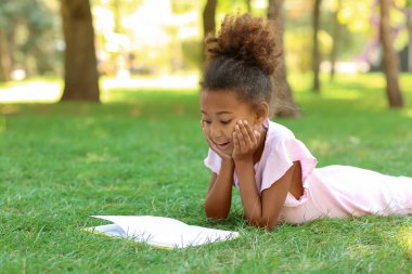 Little African-American girl reading book on grass in park