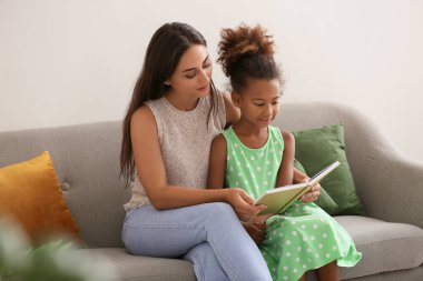 Little African-American girl reading book with her teacher on sofa