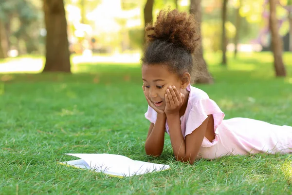 Küçük Afro-Amerikalı kız parkta çim üzerine kitap okuyor.
