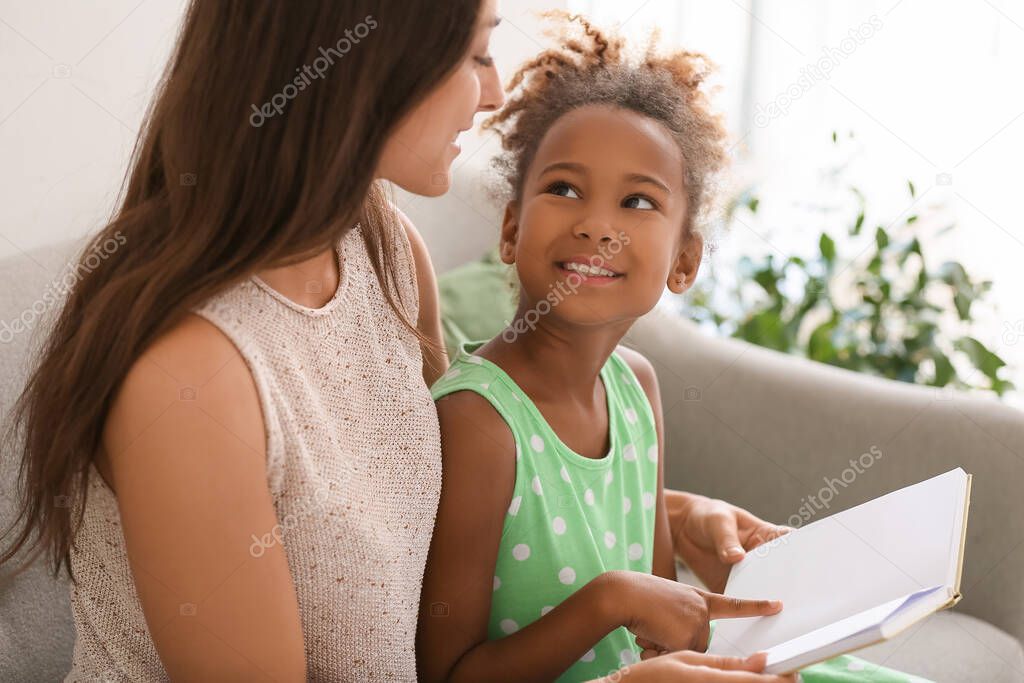 Little African-American girl reading book with her teacher on sofa