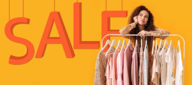 Beautiful thoughtful African-American girl near rack with clothes and word SALE on yellow background
