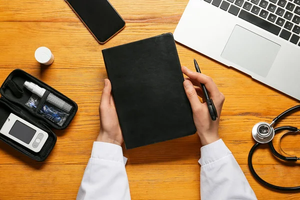 Female doctor with notebook, digital glucometer and pen on wooden background