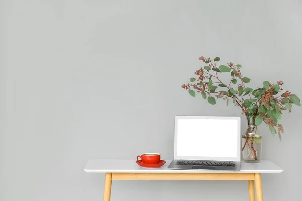 Table with laptop, plant branches in vase and cup of coffee near grey wall