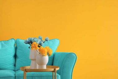 Vases with beautiful flowers on table and comfortable blue sofa near yellow wall