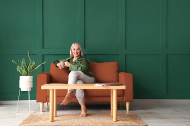 Yeşil duvarın yanında oturmuş çay içen olgun bir kadın.