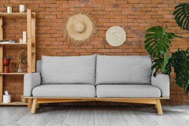 Shelving unit with decor, grey sofa and monstera plant near brick wall