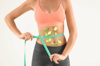 Sporty young woman with measuring tape and healthy vegetables on her body against light background. Concept of diet
