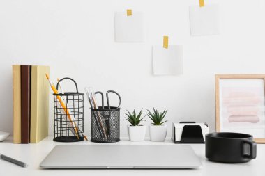 Boş notları olan bir işyeri, modern laptop, bardak ve kırtasiye malzemeleri beyaz duvarın yanında.