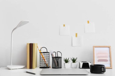 Boş notları olan işyeri, modern laptop, lamba, bardak ve kırtasiye malzemeleri beyaz duvarın yanında.