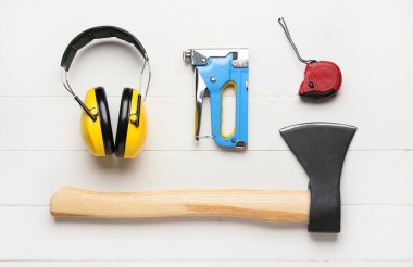 Protective yellow headphones, building stapler, ruler and ax on white wooden background