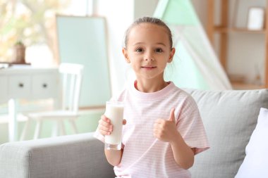 Little girl holding glass of milk and showing thumb up on sofa