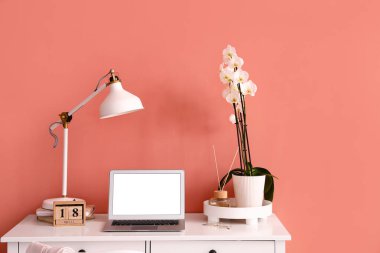 Modern laptop, lamp and beautiful orchid flower on table near color wall