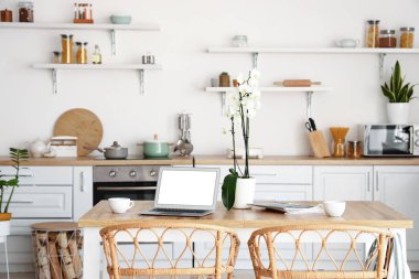 Modern laptop, fincanlar ve orkide mutfaktaki yemek masasında.