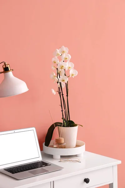 Modern laptop and orchid flower on table near color wall