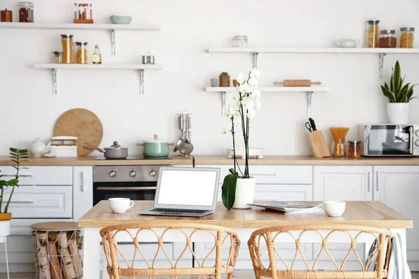 Modern laptop, fincanlar ve orkide mutfaktaki yemek masasında.