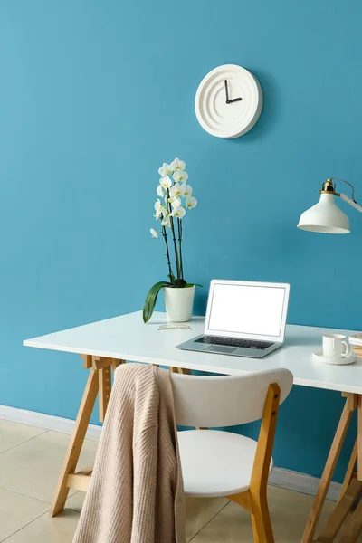 Workplace with modern laptop, orchid flower and cup near blue wall